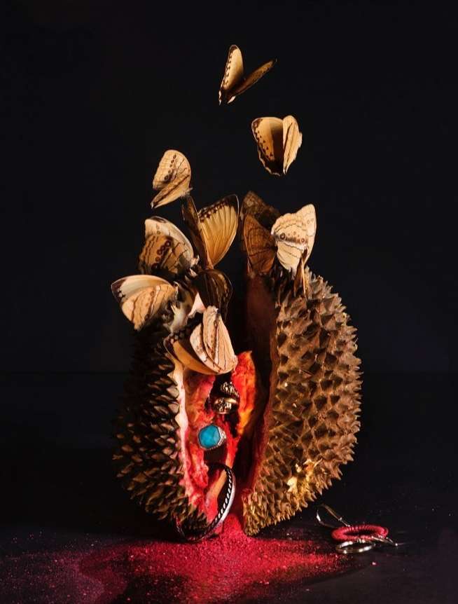 Still life shot of a halved durian with jewelry inside and butterflies emerging against a dark background with red powder details.
