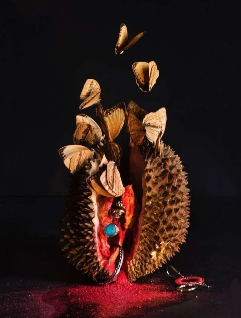 Still life shot of a halved durian with jewelry inside and butterflies emerging against a dark background with red powder details.