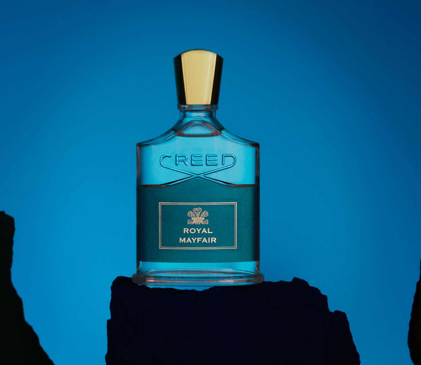 A bottle of Creed Royal Mayfair cologne sits on a textured surface against a vibrant blue background in a beauty still-life shot.