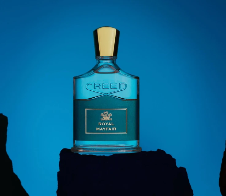 A bottle of Creed Royal Mayfair cologne sits on a textured surface against a vibrant blue background in a beauty still-life shot.