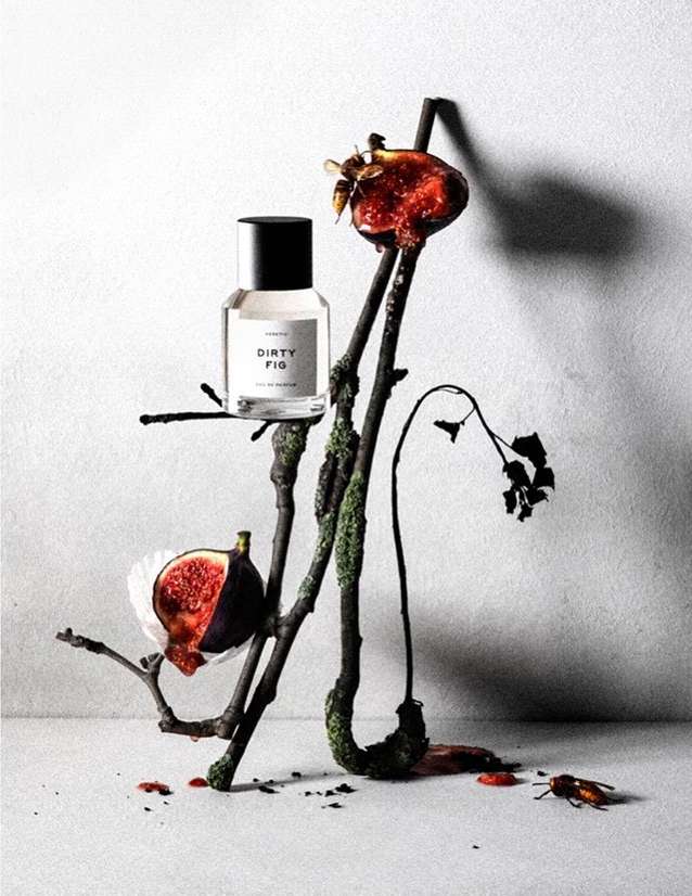 Still life of perfume bottle with figs, branches, and insects on a neutral surface against a gray backdrop in a studio setting.