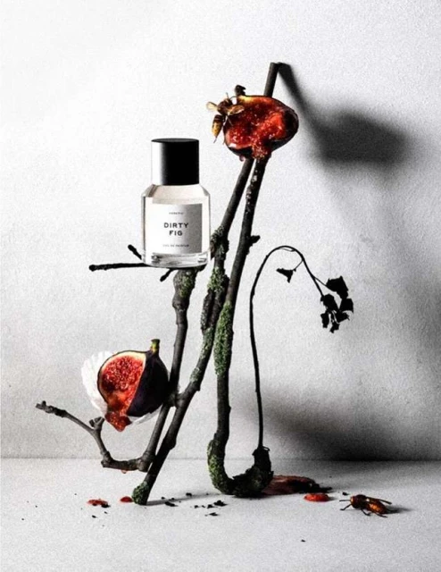 Still life of perfume bottle with figs, branches, and insects on a neutral surface against a gray backdrop in a studio setting.