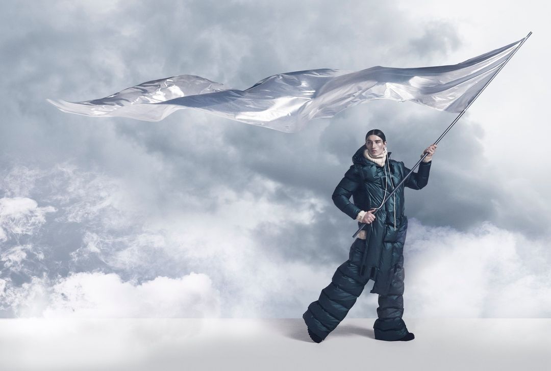 Fashion shot of a male model wearing a puffer jacket and pants, holding a silver flag against a cloudy sky backdrop in a studio.