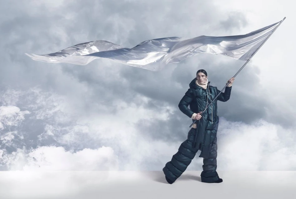 Fashion shot of a male model wearing a puffer jacket and pants, holding a silver flag against a cloudy sky backdrop in a studio.