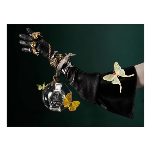 Black-gloved hand adorned with jewelry holds a Creed perfume bottle surrounded by yellow butterflies against a dark green background.