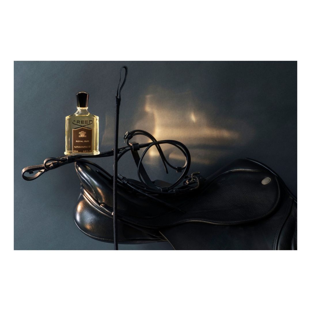 Still life of a Creed perfume bottle on a black leather saddle with a riding crop against a gray background with reflected light.