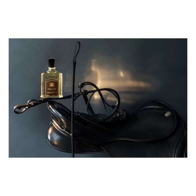 Still life of a Creed perfume bottle on a black leather saddle with a riding crop against a gray background with reflected light.