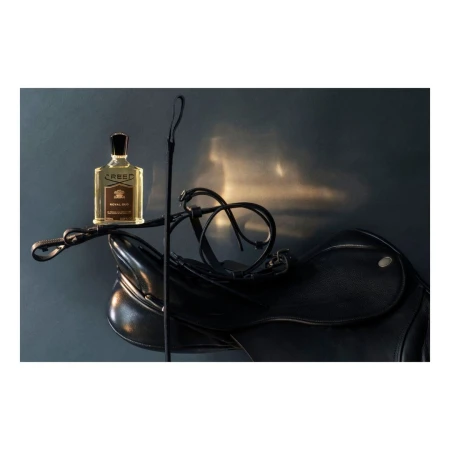 Still life of a Creed perfume bottle on a black leather saddle with a riding crop against a gray background with reflected light.