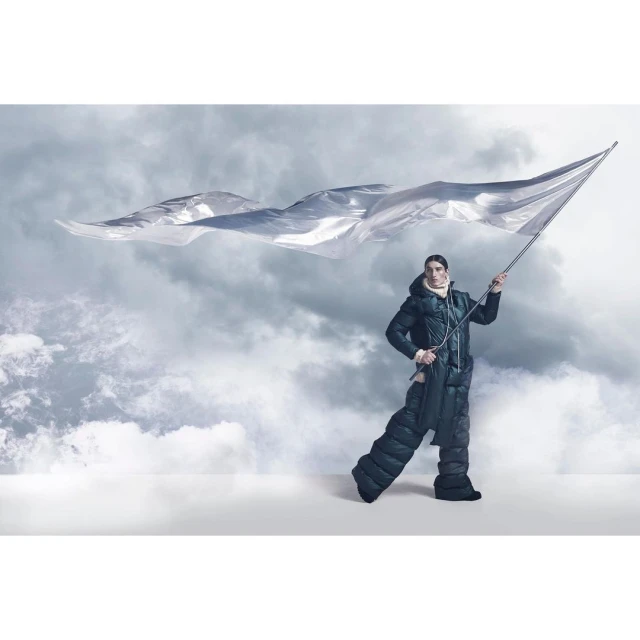 A man in a puffy coat and matching pants holds a waving silver flag against a cloudy sky backdrop in a fashion editorial shot.
