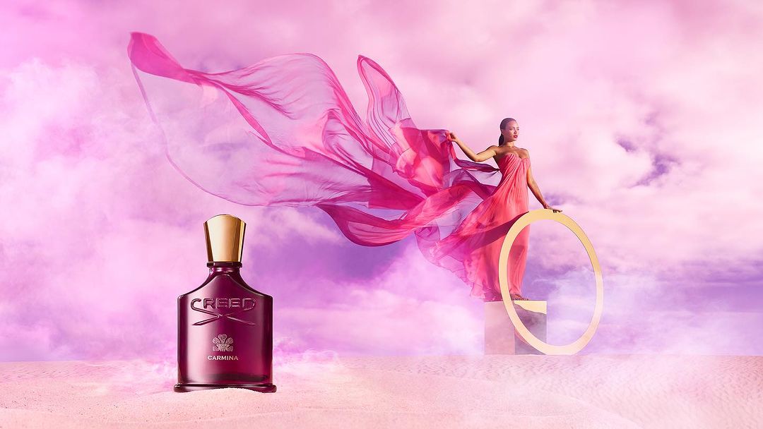 Creed Carmina perfume bottle with woman in pink dress and flowing fabric against a pink sky backdrop, beauty and fashion.