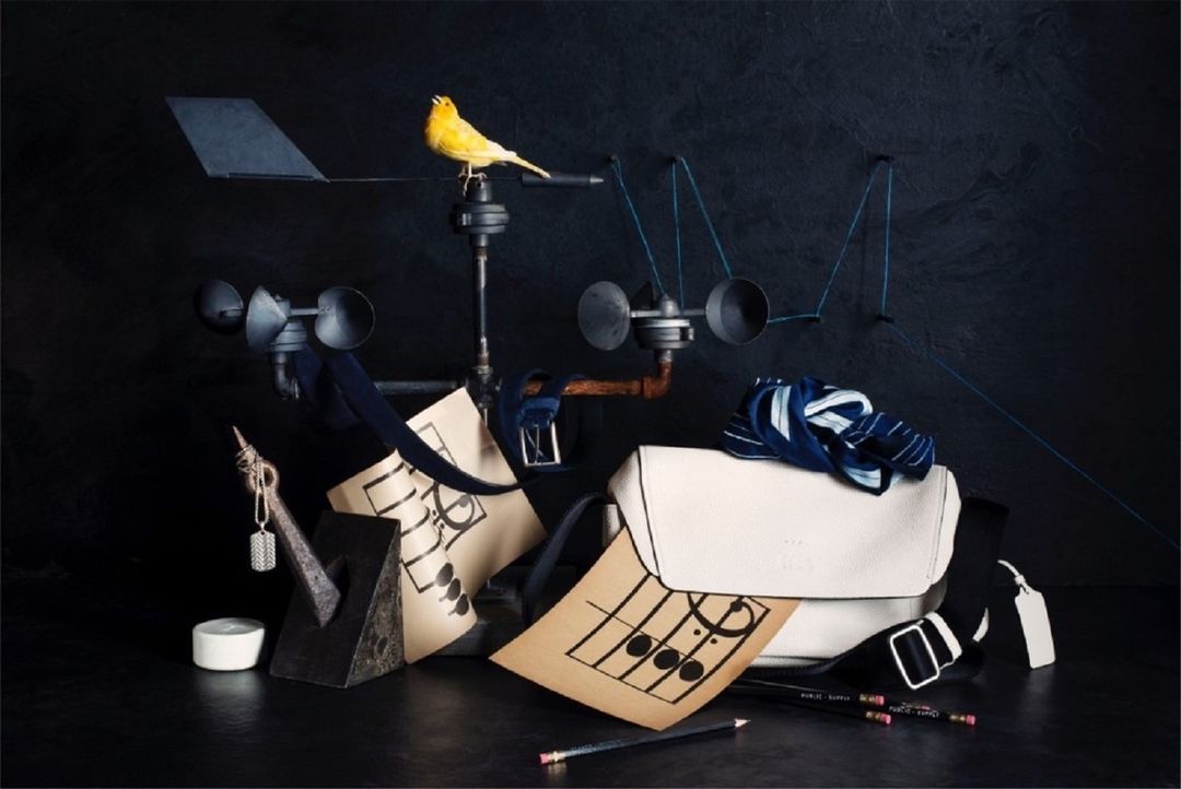 Still life photo with a yellow bird perched atop a vintage anemometer, accompanied by a white satchel, pencils, and sheet music.