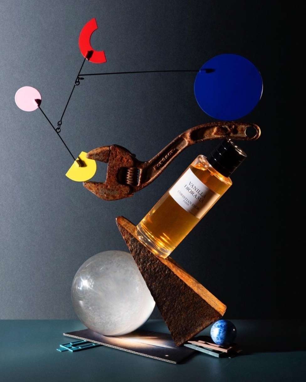 Product shot of perfume bottle balanced with geometric objects, a wrench, and crystal ball in a dark studio with dramatic lighting.