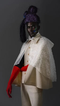Person with dark skin and vibrant hair wearing a textured suit and bubble wrap cape with bright red gloves against a gray backdrop.