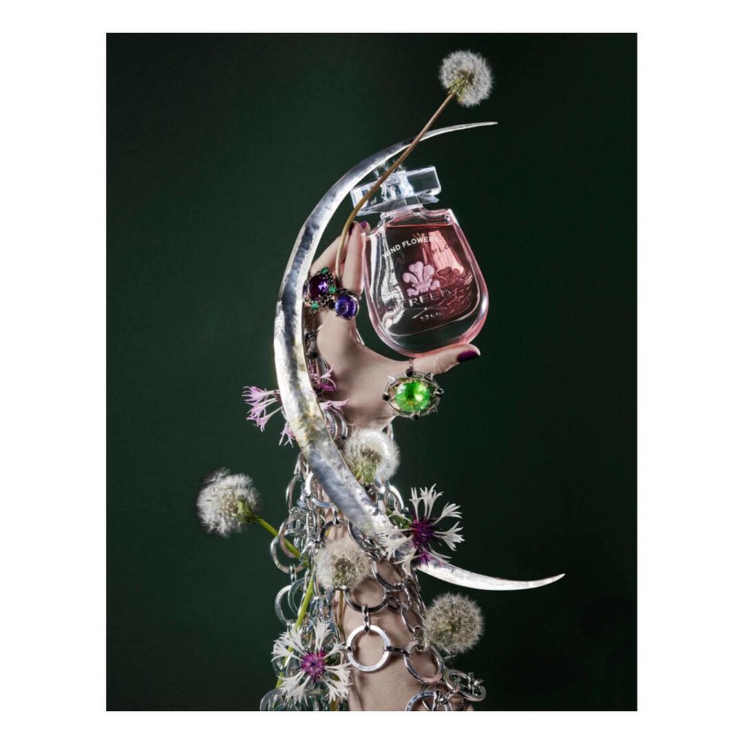 Still life of a pink perfume bottle held by a hand adorned with rings and silver bracelets, set against a dark green backdrop with dandelions.
