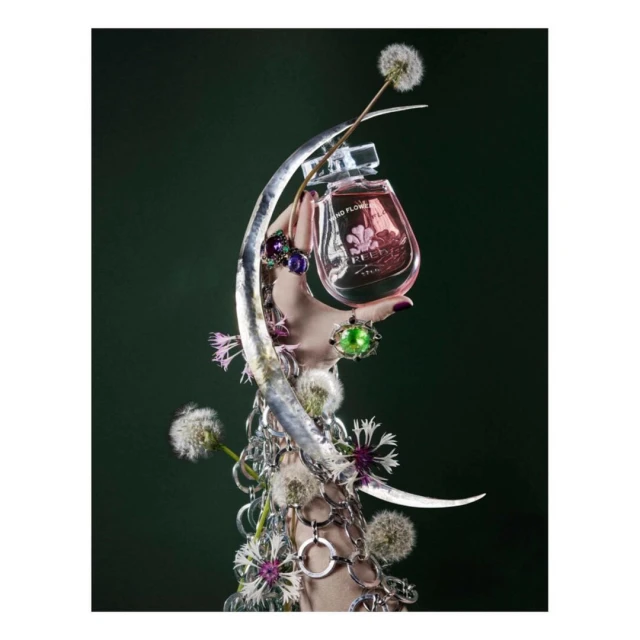Still life of a pink perfume bottle held by a hand adorned with rings and silver bracelets, set against a dark green backdrop with dandelions.
