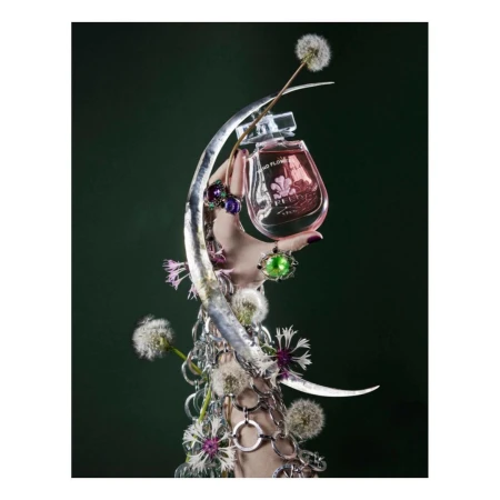 Still life of a pink perfume bottle held by a hand adorned with rings and silver bracelets, set against a dark green backdrop with dandelions.