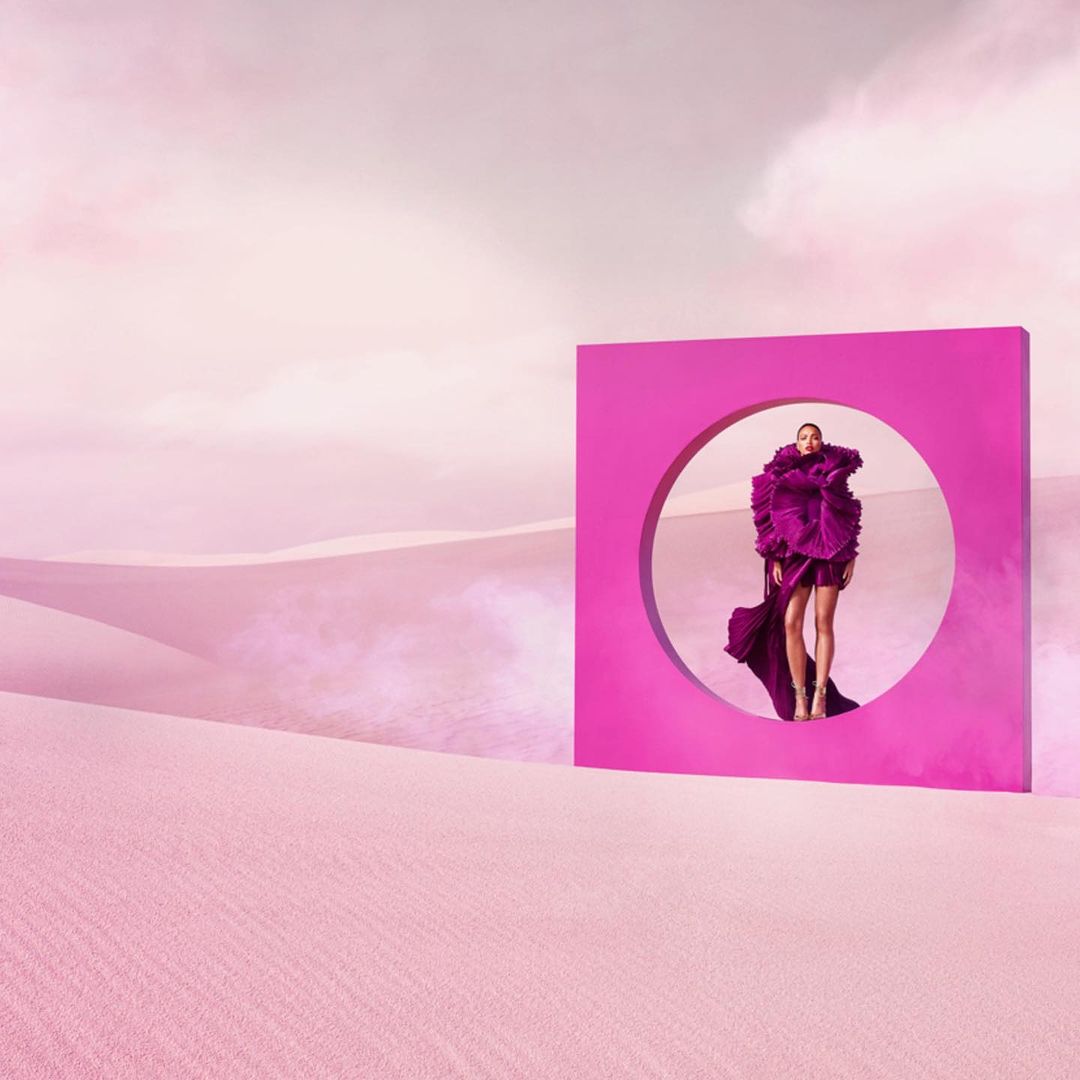 Woman in magenta gown standing inside a magenta square frame with a circular cut-out, set in a pink desert landscape under a cloudy sky.