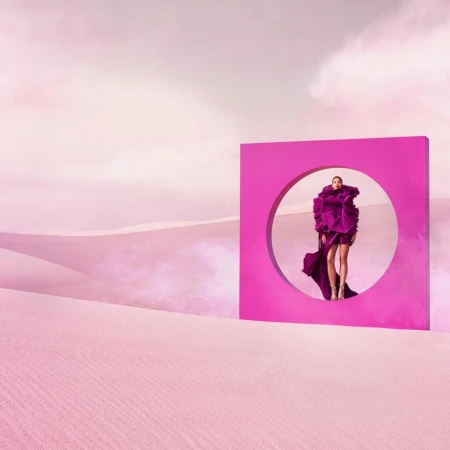 Woman in magenta gown standing inside a magenta square frame with a circular cut-out, set in a pink desert landscape under a cloudy sky.