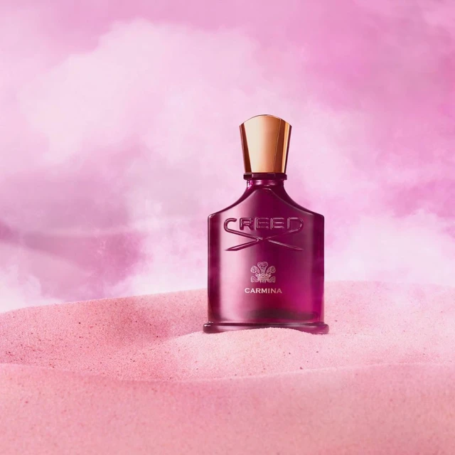 Creed Carmina perfume bottle stands prominently on pink sand, set against a softly blurred pink and white sky background.