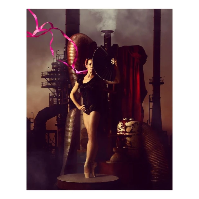 A ballerina stands on a platform with a ribbon and hand fan in front of industrial machinery and smokestacks in a stylized artistic photograph.