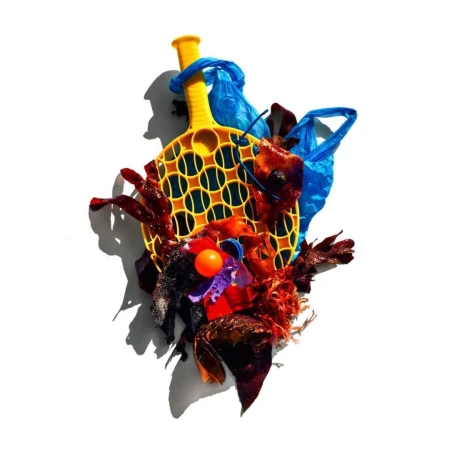Abstract still life of a yellow paddle with an orange ball, decorated with blue plastic bags and red and brown abstract objects on white.