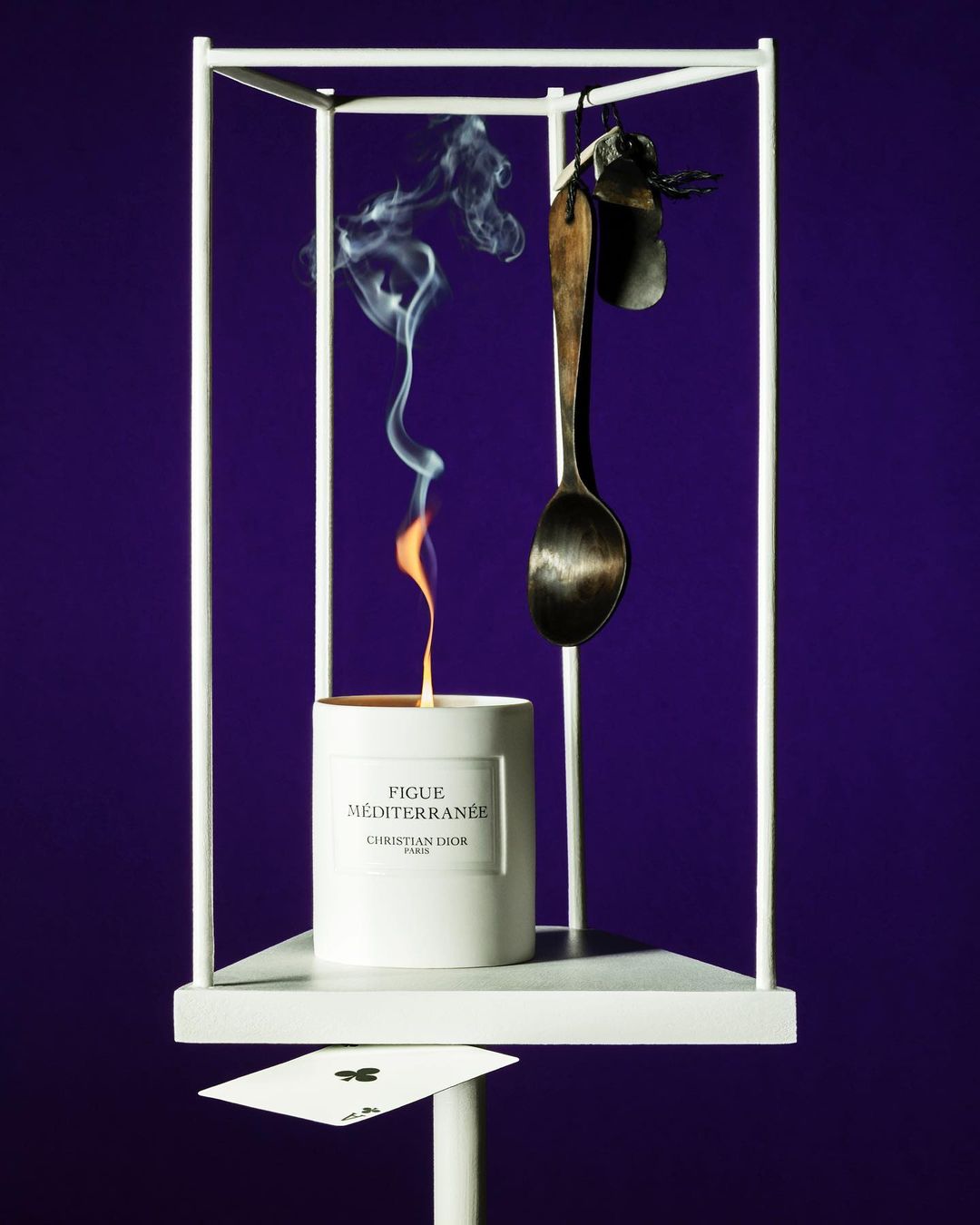 A candle and vintage spoon hang in a white display case against a purple background. A playing card is underneath on a white stand.