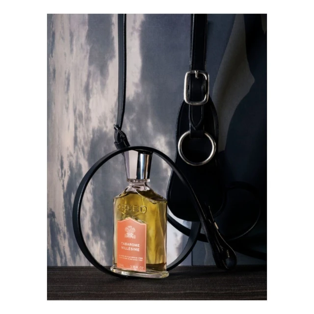 Glass bottle of Creed Tabarome Millesime fragrance in a black metal ring, paired with black equestrian tack, on a grey textured surface.