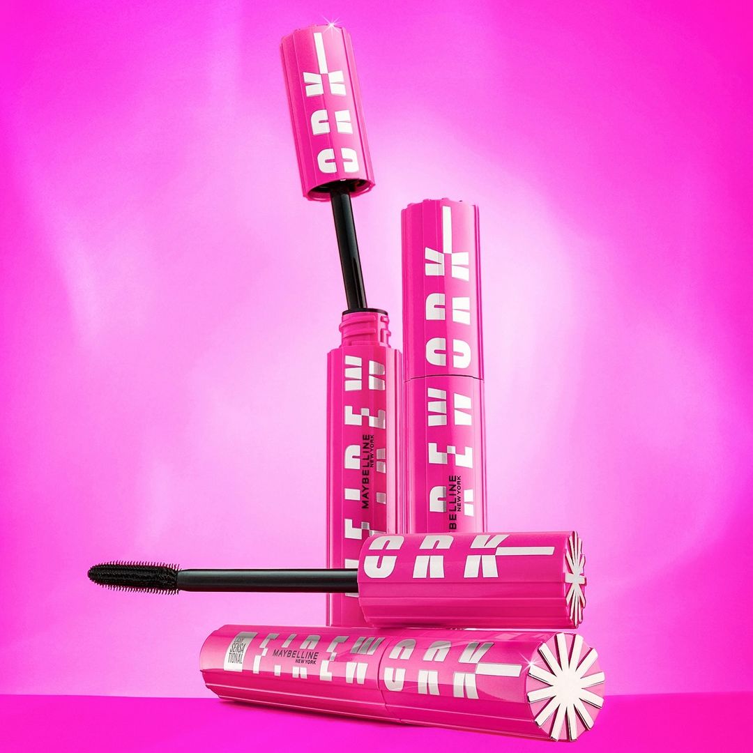 Product shot of Maybelline Firework mascara tubes in pink packaging against a matching pink gradient background with studio lighting.