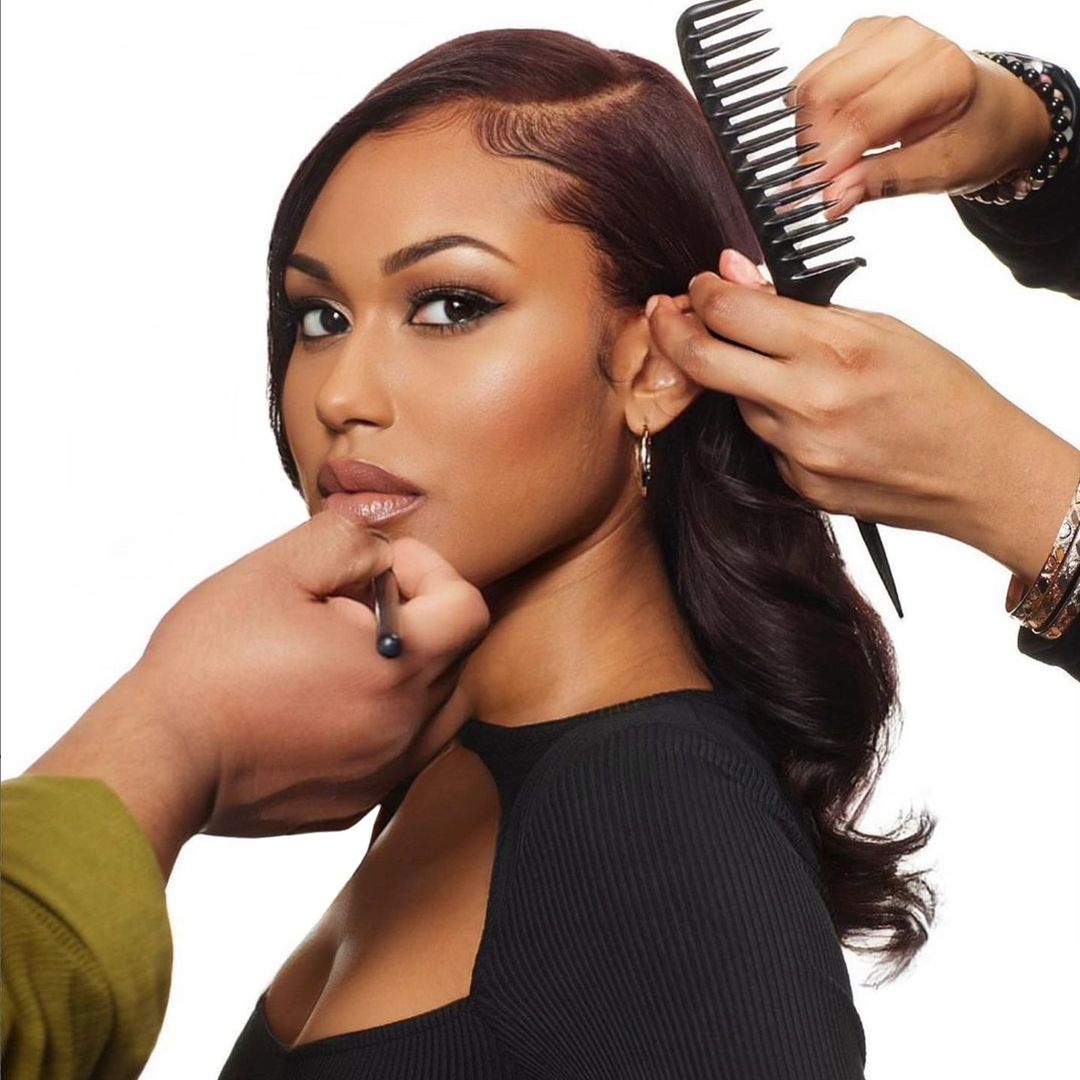 Close-up of a Black woman getting her hair styled with a comb and makeup applied to her face by two stylists in a studio setting.
