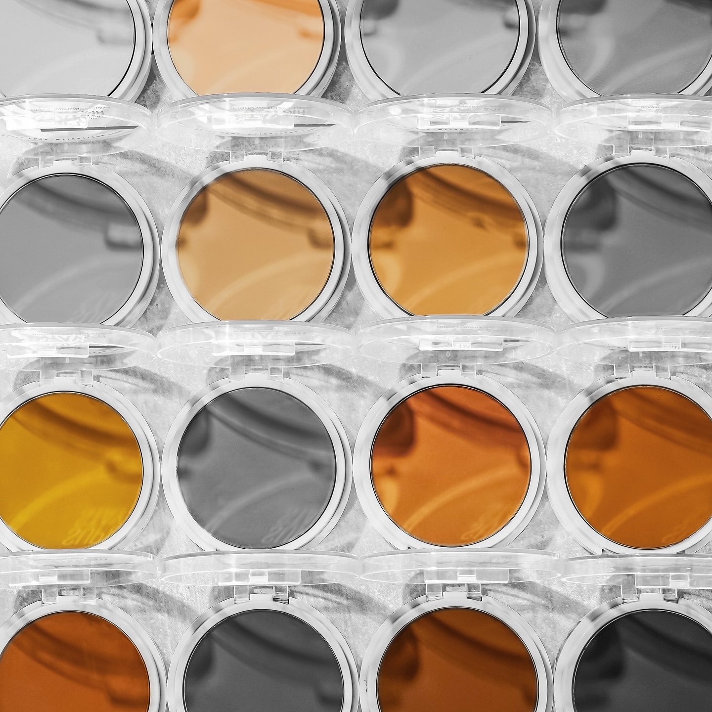Close-up of an array of powder compacts in a grid pattern, featuring a color palette of oranges, browns, and grays in a studio setting.