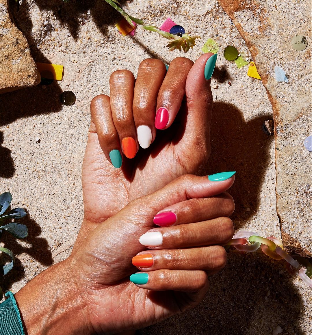 Close-up of manicured hands with multicolored nail polish on a sandy surface with confetti and plants, illuminated by sunlight.