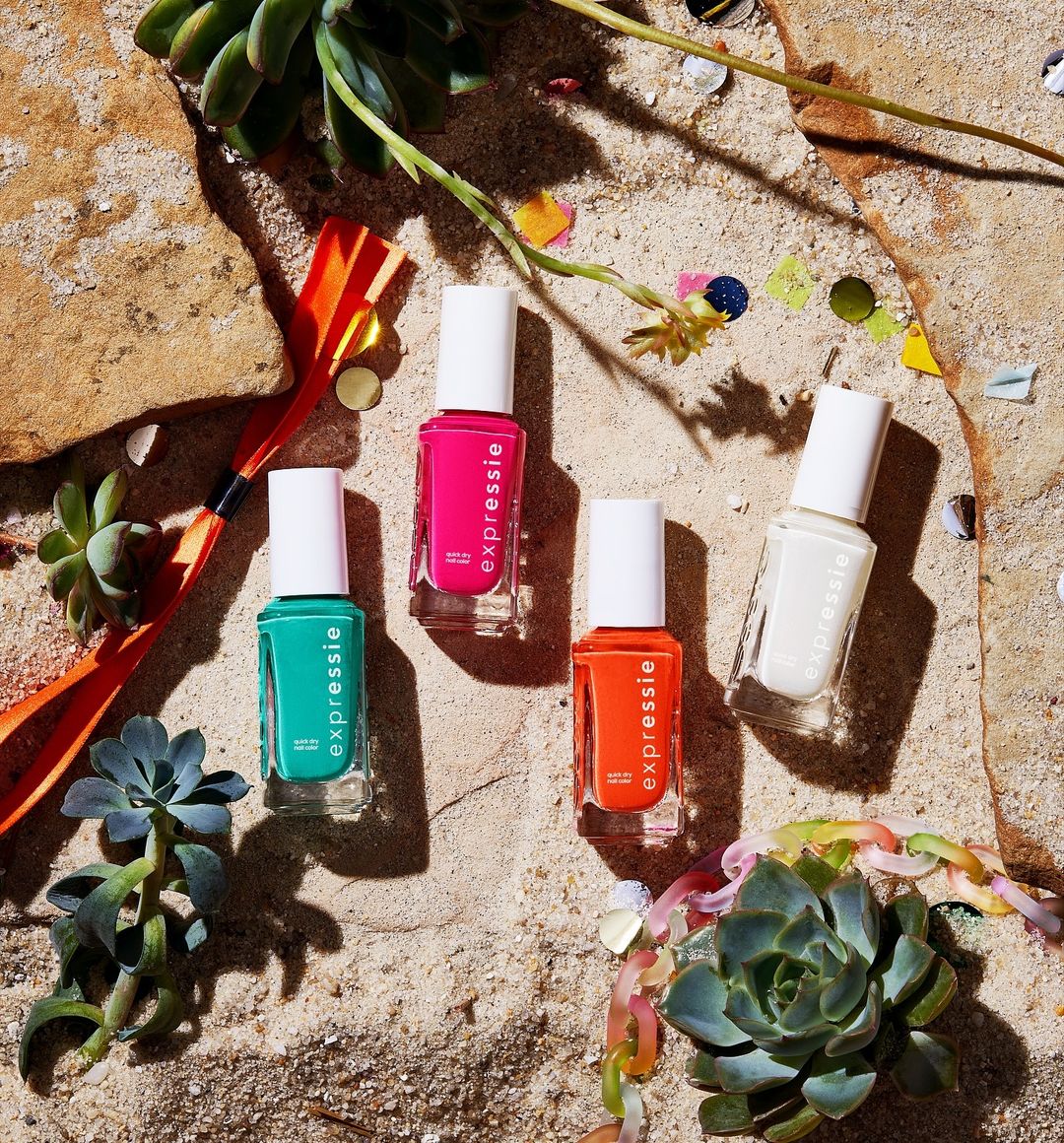 Close-up of four Essie nail polish bottles in pink, orange, white, and teal, arranged on a sandy surface with confetti and succulent plants.