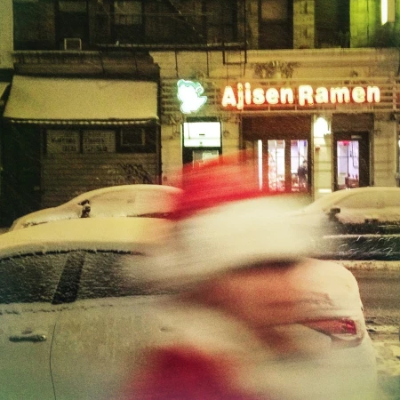 Snow-covered cars line a city street at night near a brightly lit ramen restaurant in a nighttime urban winter scene.