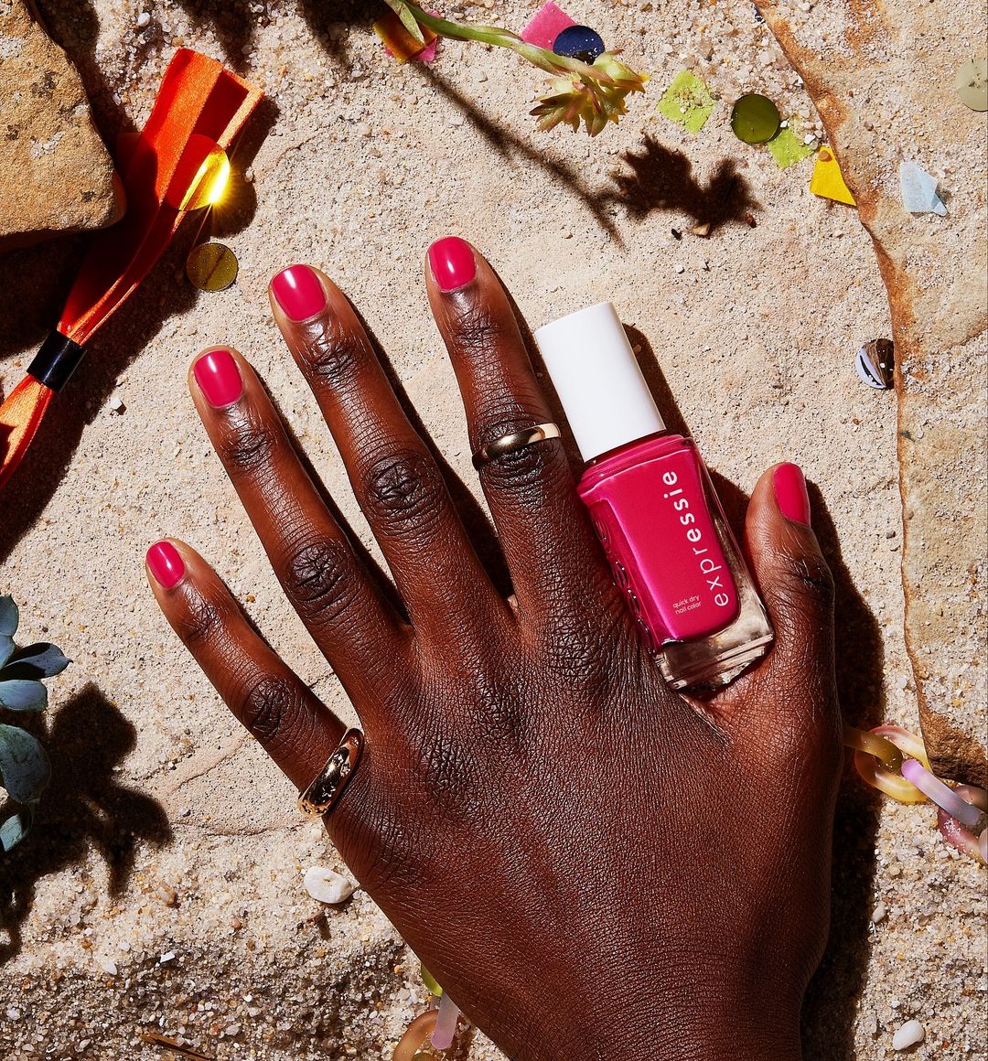 Close-up of a dark-skinned hand with pink polished nails holding an Essie Expressie nail polish bottle on a sandy surface.