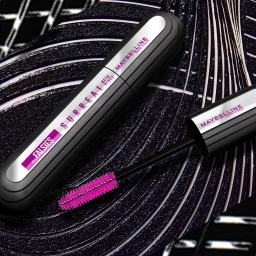 Maybelline The Falsies Surreal mascara tube and open wand with vibrant pink bristles resting on a textured dark patterned surface.