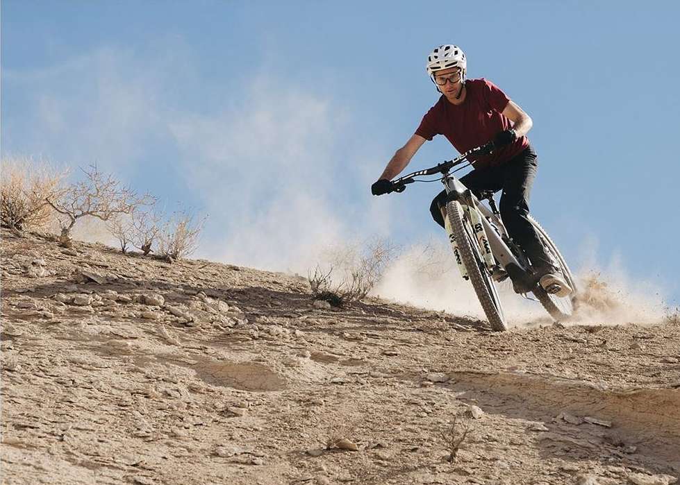 Adult man mountain biking downhill on a dirt trail wearing helmet, sunglasses, red shirt and black pants with a blue sky overhead.