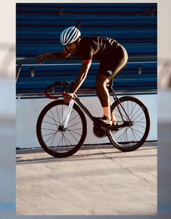 Adult man in cycling helmet riding a black bicycle on a velodrome track, wearing a dark athletic shirt and shorts, captured in daylight.