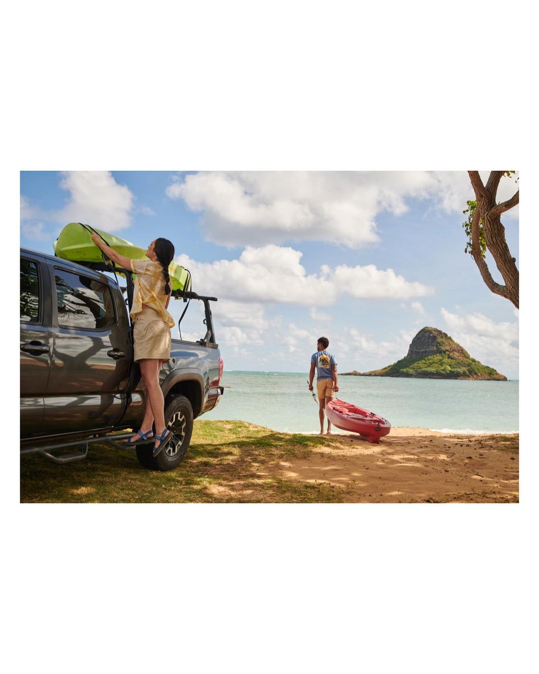 A woman standing on the step of a pick up truck taking a kayak off the roof rack while another person pulls a kayak on a beach.