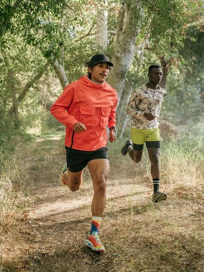 Two men running on a dirt path surrounded by trees in an outdoor environment. They are wearing athletic apparel and running shoes.