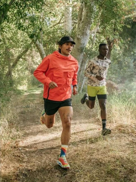 Two men running on a dirt path surrounded by trees in an outdoor environment. They are wearing athletic apparel and running shoes.