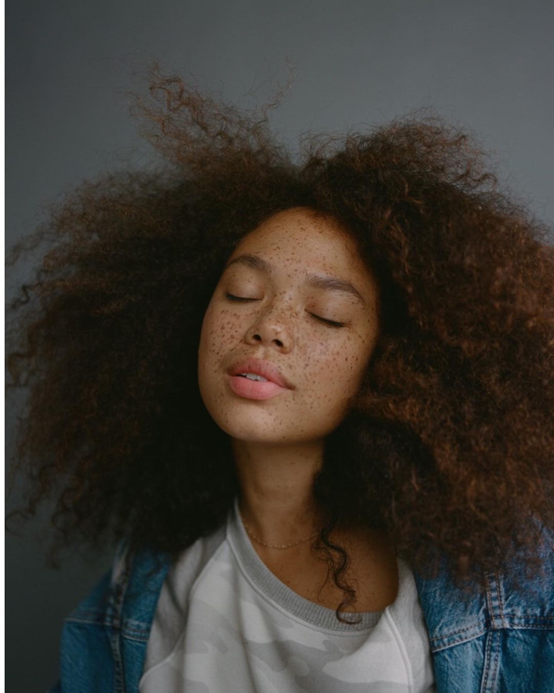 Portrait of a young freckled woman with curly dark hair, eyes closed, wearing a camo-patterned shirt and denim jacket against a gray background.