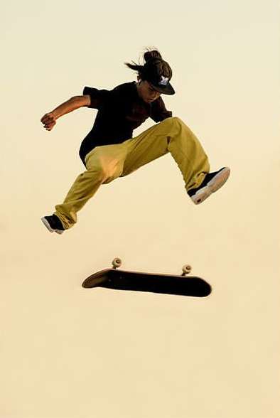 Teen skater in mid-air performing a jump with a skateboard against a clear sky, showcasing athleticism and skill in action sports.