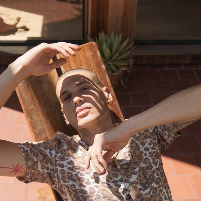 An adult male with short hair poses in a printed shirt on a wooden chair outside with light shadows on his face and arms.