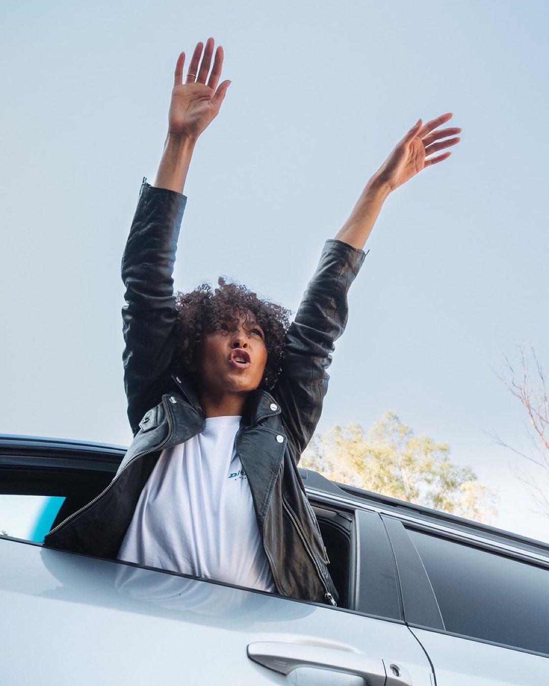 Woman with curly hair wearing a black leather jacket raising her arms out of a car window