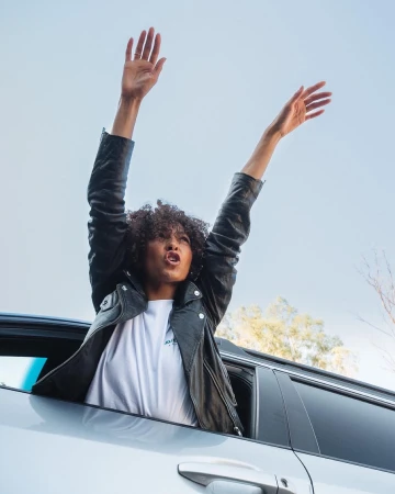 Woman with curly hair wearing a black leather jacket raising her arms out of a car window