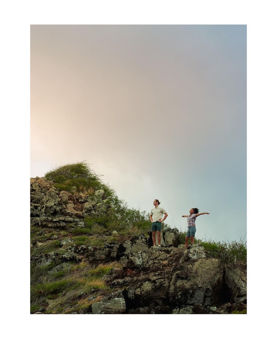 A couple stands atop a rocky hill overlooking a scenic landscape with a cloudy sky. They are enjoying the natural beauty around them.