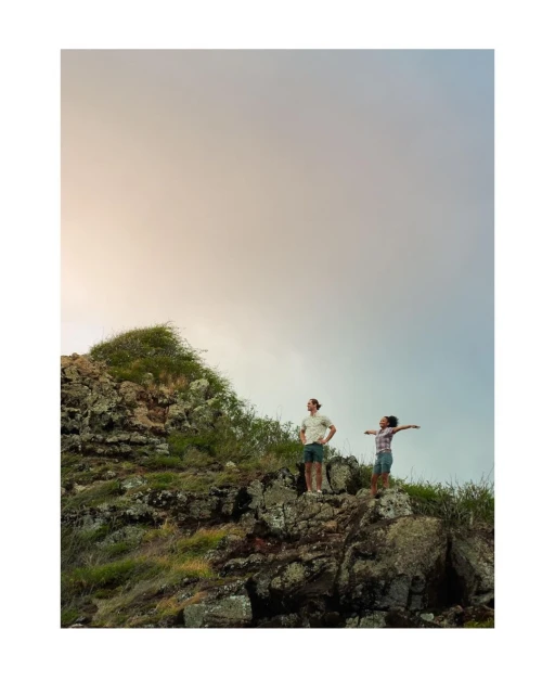 A couple stands atop a rocky hill overlooking a scenic landscape with a cloudy sky. They are enjoying the natural beauty around them.