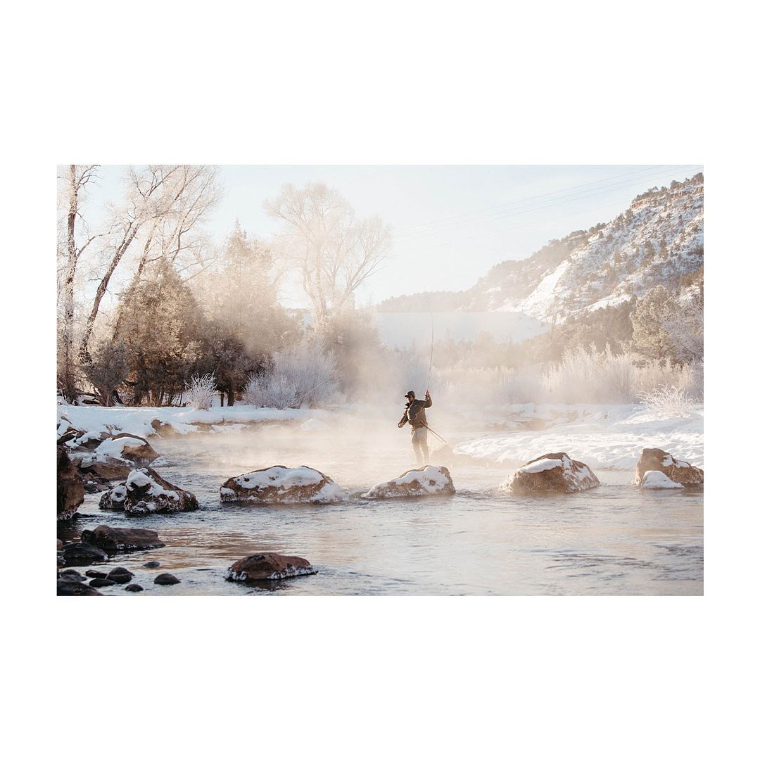 Fly fisherman wading across a stream on rocks in the winter snow near frosted trees with a bright sky.
