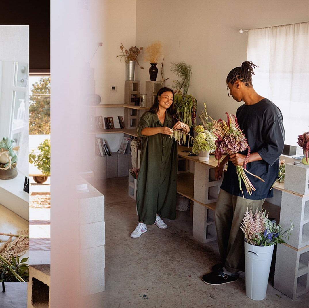Two adults arranging flowers in a bright flower shop. Woman trims stems while man holds a bouquet in a room filled with shelves.