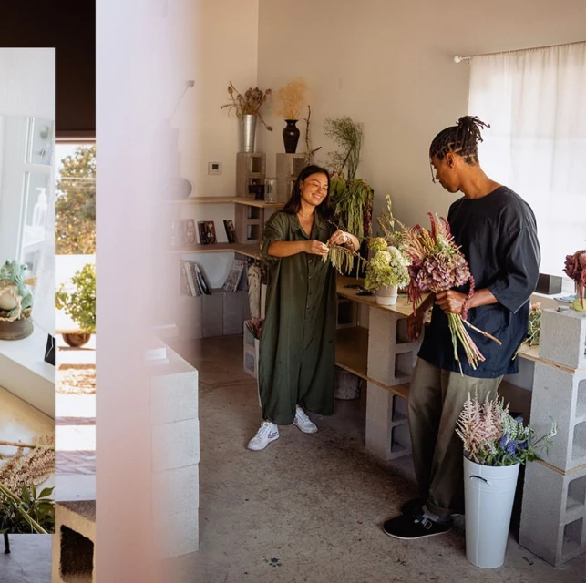 Two adults arranging flowers in a bright flower shop. Woman trims stems while man holds a bouquet in a room filled with shelves.